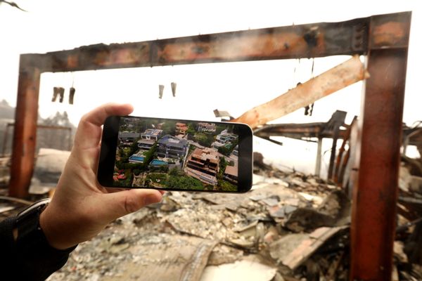 A hand holding a phone showing a large home. Behind the hand and phone is the ruins of that home after a fire. 