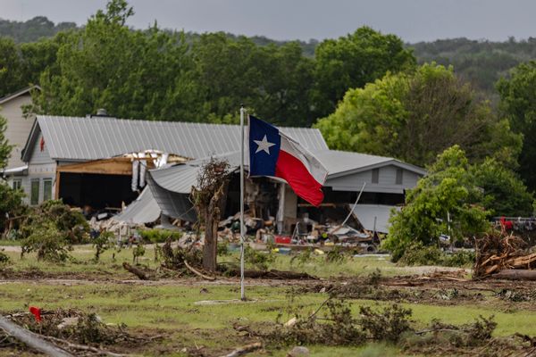 A blue, white and red striped flag with a single star — the Texas flag — in front of a partially destroyed house. 