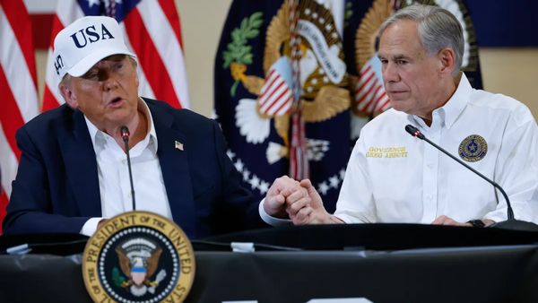 Two white men sit next to each other. The man on the left wears a blue suit and white hat with blue letters that say USA, while the man on the right wears a white button-down.