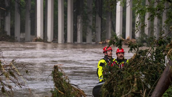 Two rescue workers in bright yellow and black gear with red helmets navigate floodwaters on an inflatable boat. They are surrounded by tree branches and debris, with a bridge supported by tall concrete columns in the background. 