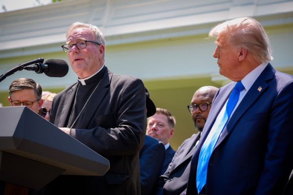 U.S. President Donald Trump, a white man with a tan, looks at a white man in a white clerical collar and black clergy robe. 