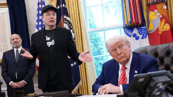 Elon Musk stands and gestures while speaking in the Oval Office, wearing a black jacket, black shirt and a cap. President Donald Trump sits at the Resolute Desk looking to his left toward Musk. Another man in a suit stands in the background. American and military flags and gold curtains are visible behind them.