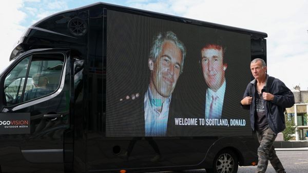 A person passes by a truck that shows two white men smiling. At the bottom, white text says, "WELCOME TO SCOTLAND, DONALD."