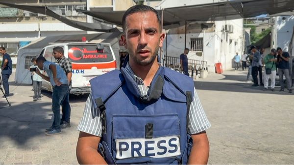A male journalist wearing a blue press vest stands in the foreground, looking directly at the camera. Behind him, several people, including other journalists and bystanders, are gathered near an ambulance marked with "AMBULANCE" and "M.O.H" in both English and Arabic. The scene takes place outdoors in what appears to be a medical or emergency facility, with white buildings and some shade overhead. 