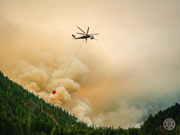 A helicopter with a bucket hanging from below flies over a smoky forest. 