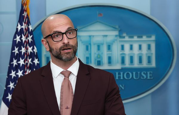 A bald white man with a short cropped salt and pepper beard, black rectangular glasses and a pink tie speaks at the White House podium. 