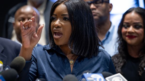 Close up of Jasmine Crockett speaking in front of microphones. Her hand is raised and there are people in the background behind her