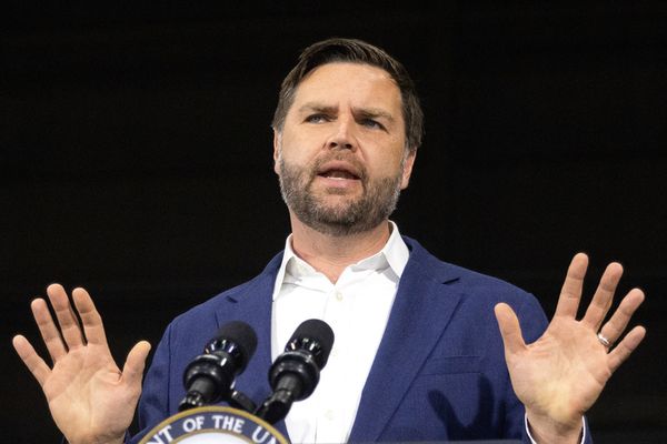 A white man with short cropped brown hair and a beard, U.S. President JD Vance, holds his hands up while speaking in front of a microphone. 