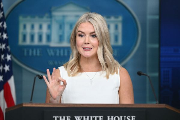 A white woman with shoulder-length blonde hair wearing a cross necklace and a white dress does an "OK" sign with her hands while she talks in front of a White House podium.