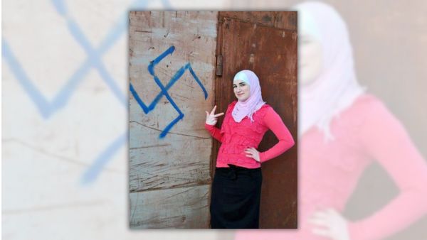 Image of woman wearing a pink hijab posing next to a wood wall with a swastika in blue graffiti.