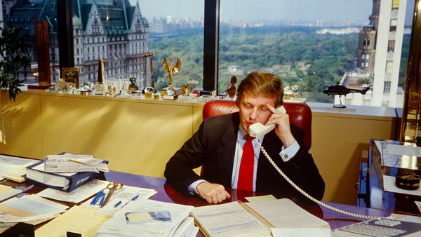 In this image from the 1980s, a white man sits at a desk with papers scattered across it. He is holding a white analog phone up to his ear. The window behind him overlooks multiple buildings.