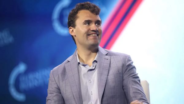 A white man smiles while looking to the right. He is sitting down and wearing a gray suit.