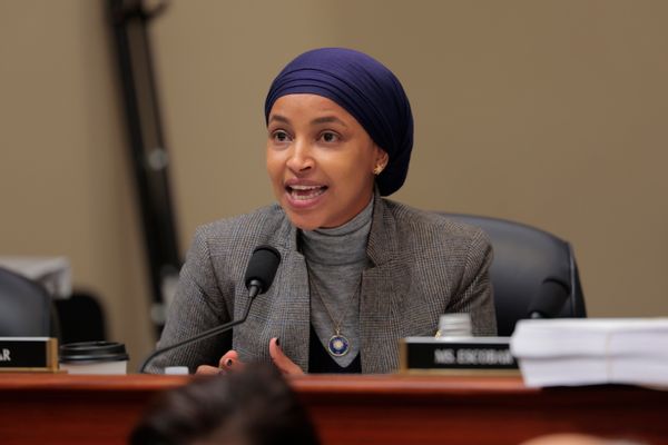 A Somali American woman sitting at a desk wearing a gray pinstriped suit and a purple head wrap covering her hair. 