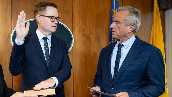 On the left, a middle-aged white man with short hair wearing glasses and a suit holds up one hand and keeps another on a book being held up by someone to the left of the camera. An older white man with short greying hair, also in a suit, speaks to the man on the left.