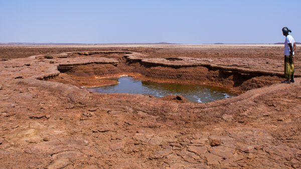 Photo of man at the edge of an acid pond cratered into reddish desert surface, sky is clear and blue above