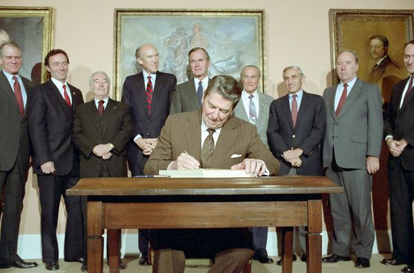 A vintage photo from the 1980s shows President Ronald Reagan, a white man with coiffed hair in a brown suit, signing a stack of papers while surrounded by other white men in suits.