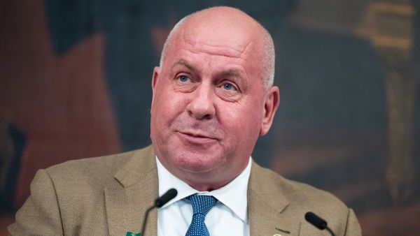 A man with a bald head and light complexion wears a tan suit jacket, white dress shirt and blue patterned tie. He is speaking into a microphone. The background is blurred, possibly showing a mural or painting.