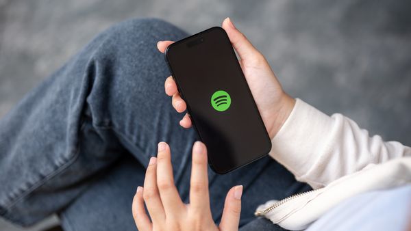 Person wearing jeans and white sweatshirt is shown holding a black cellphone displaying the green Spotify logo