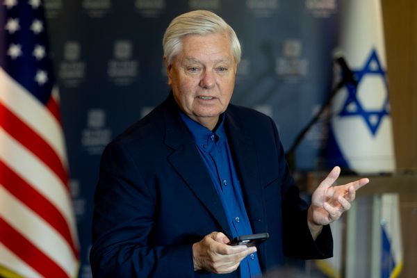 U.S. Senator Lindsey Graham, a white man with combed back short white hair, stands in front of U.S. and Israel flags. 