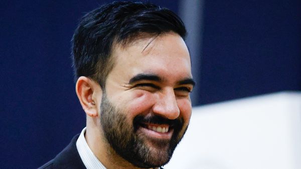 A South Asian man wearing a dark suit smiles widely.
