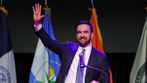 Zohran Mamdani, wearing a suit and tie, stands at a podium and waves with his right hand. Several flags are displayed vertically behind him.