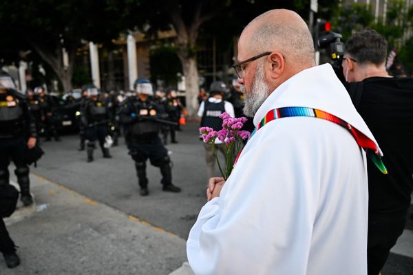 A priest wearing white robes with a colorful stole holds a bunch of purple flowers. He is standing with his head bowed in front of law enforcement wearing heavy tactical gear. 