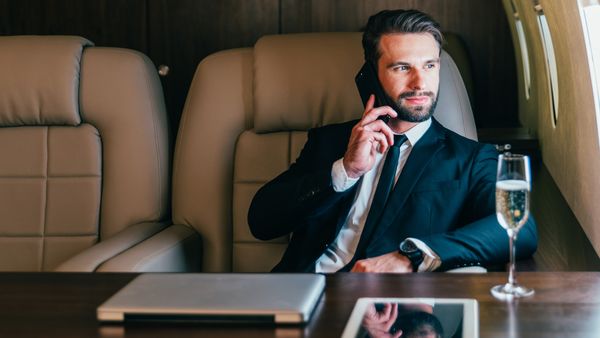 A well-groomed white man with a mustache and beard wearing a dark suit sitting on a large chair inside a private jet, holding a phone up to his ear. 