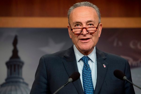 Chuck Schumer, a white man wearing a dark blue suit and red glasses halfway down his nose, speaks at a podium.