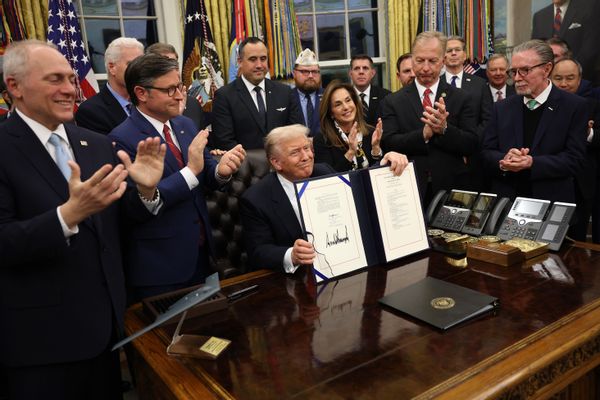 U.S. President Donald Trump holds up a signed bill and smiles sitting behind a desk in the Oval Office. People surrounding him clap. 