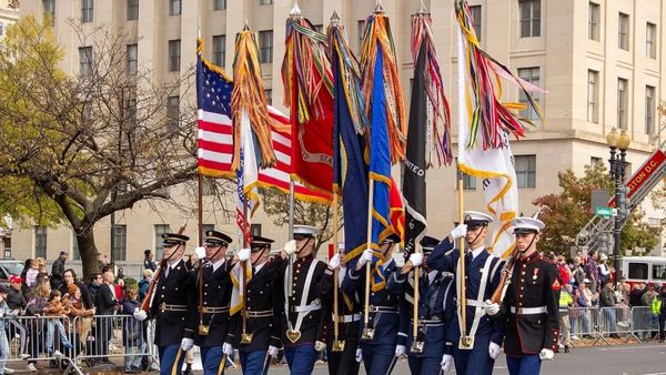 A line of people wearing military uniforms hold flags while walking in a parade. People stand to the side watching them.