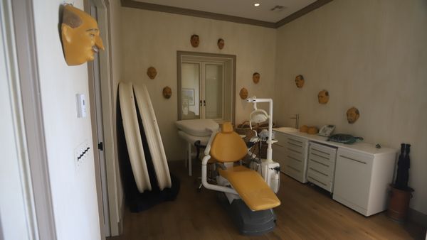 A picture of a room showing masks of older men along the walls, many of whom are balding. In the middle of the room sits what appears to be a dentist's chair.