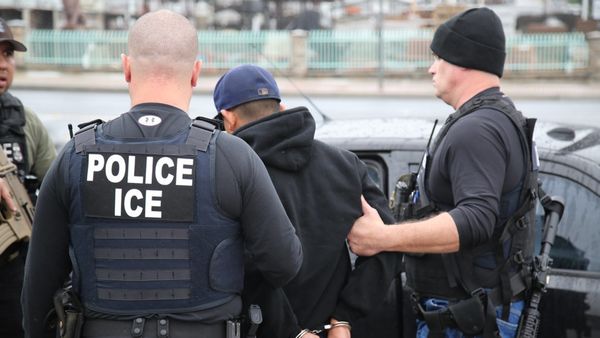 Two ICE officers escort a handcuffed man into a car. 