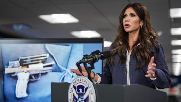 Department of Homeland Security Secretary Kristi Noem, a brunette white woman, stands in front of a screen showing a pistol.