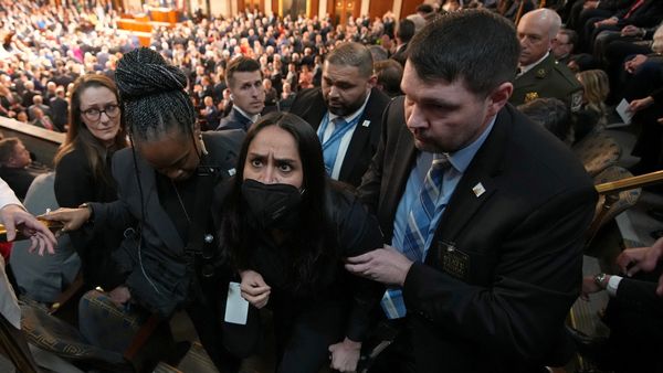 Two officers in suits hold the arms of Aliya Rahman, a Bangladeshi American woman wearing a black mask, amid a large audience in a congressional gallery. 