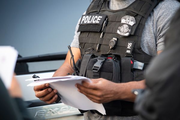 An ICE officer wearing a badge and vest holds a piece of paper. 