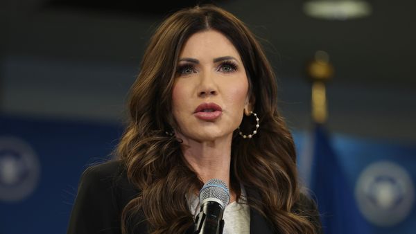 Close up photo of Kristi Noem, a white woman, from the chest up. A microphone is in front of her and a backdrop has blurry DHS seals