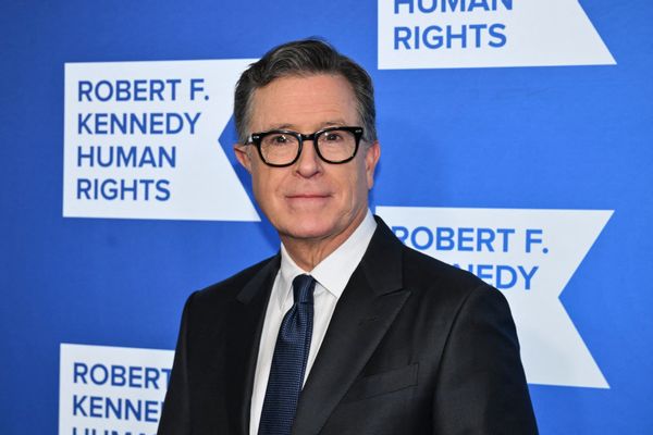 Late show host Stephen Colbert, a white man with dark hair and glasses, wearing a suit and tie