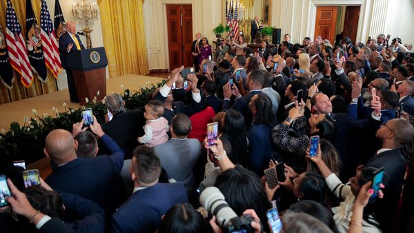 A white man stands behind a podium on a stage. A large crowd can be seen standing in front of him, many filming the man.