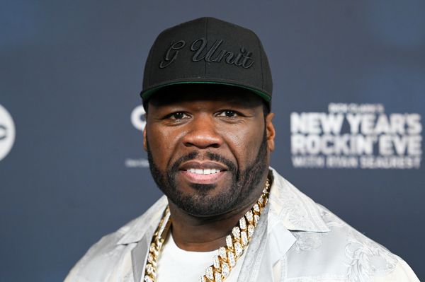 50 Cent, a Black man with a short beard, smiles while wearing a a white collared shirt. He also wears an all-black hat reading "G Unit" and a large gold chain.