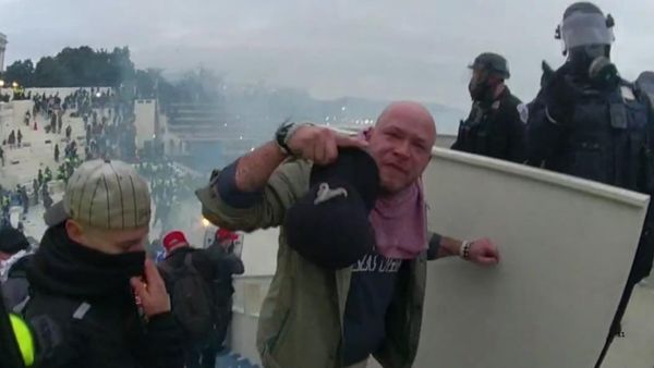 A screenshot from body camera footage shows a bald white man appearing to gesture at the person behind the body camera while holding a hat and standing on a U.S. Capitol building with a crowd behind him. 