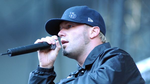 Musician Fred Durst, a white man with blonde hair and sideburns, wears a baseball hat emblazoned with "FD" and sings into a microphone.