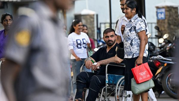 Image shows injured man in black shirt and pants in a wheelchair surrounded by other people
