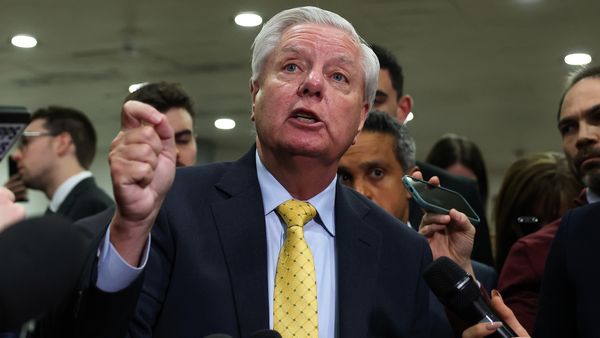 Sen. Lindsey Graham, an older white man with short white hair in a yellow tie, stands among a throng of reporters and speaks.