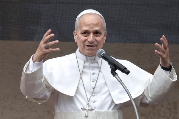 Pope Leo XIV, an older white man with glasses wearing a white frock and simple, papal hat, raises his hands as he speaks into a microphone.