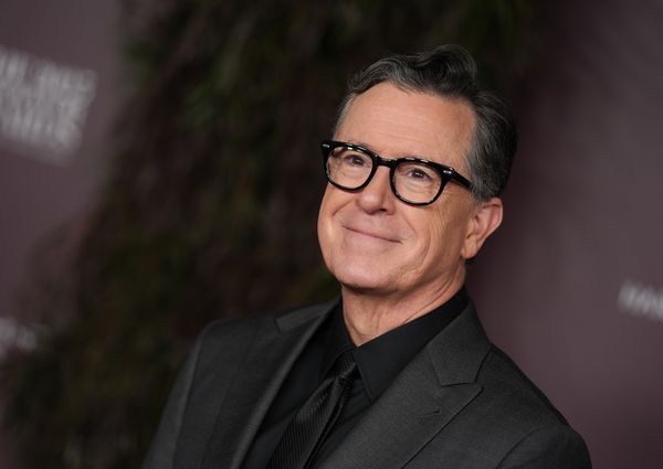 Stephen Colbert, a white man with dark, slicked back hair and glasses, wearing an all-black suit.