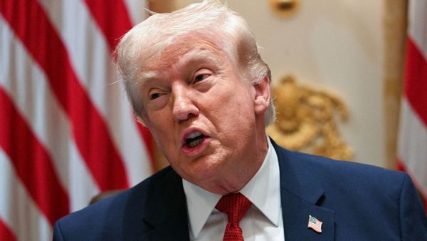 U.S. President Donald Trump, a white man wearing a suit, appears to be talking while in front of a background featuring American flags.