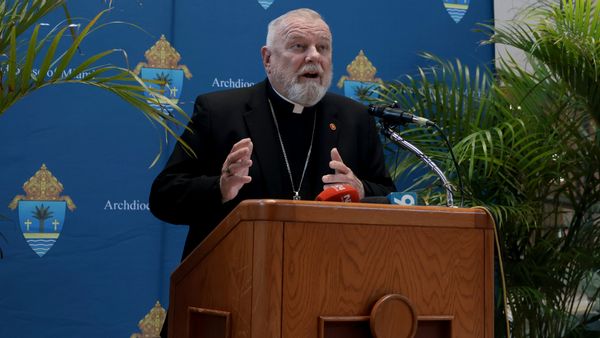 A photo of Archbishop Thomas Wenski, an older white man with white hair, beard, and moustache, speaking into a microphone on a lectern. Behind him is a backdrop featuring the Archdiocese of Miami name and logo