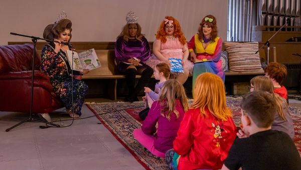 Children sit on a rug while a drag queen reads from a picture book and three others sit on a nearby bench.