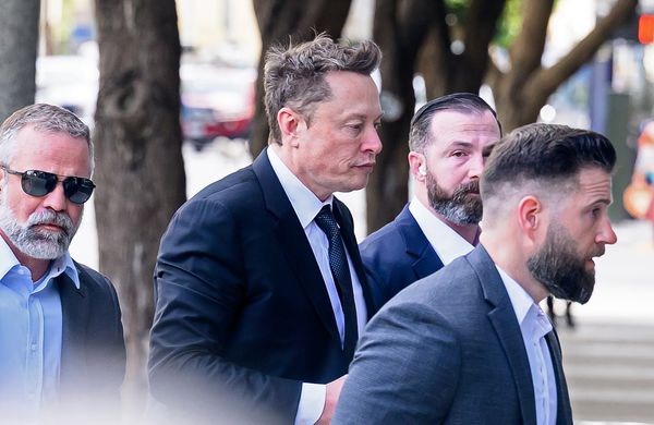 Elon Musk, a middle-aged white man with a slightly greying quiff, walks while surrounded by a posse of three men.