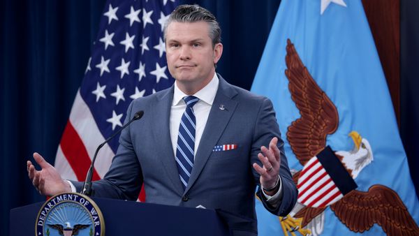 Defense Secretary Pete Hegseth, a white man with slicked back salt and pepper hair in a suit, stands in front of a podium and holds his hands in a shrug gesture.